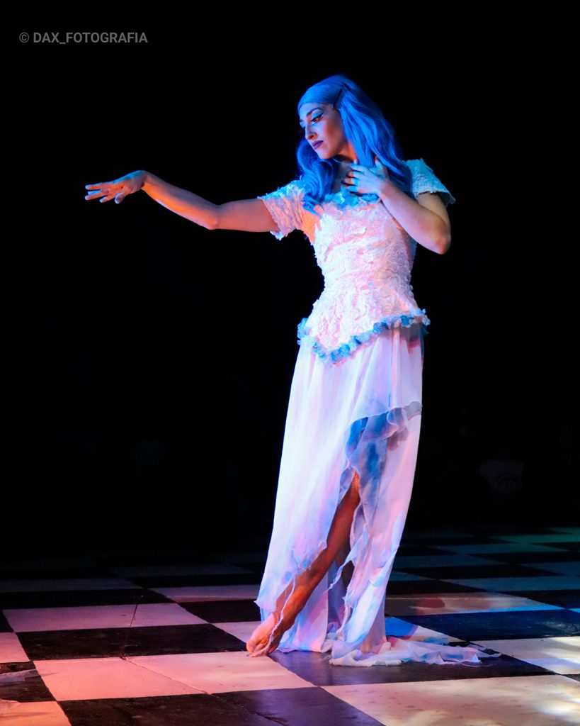 Intérprete con vestido blanco extiende los brazos en un escenario oscuro iluminado en azul.