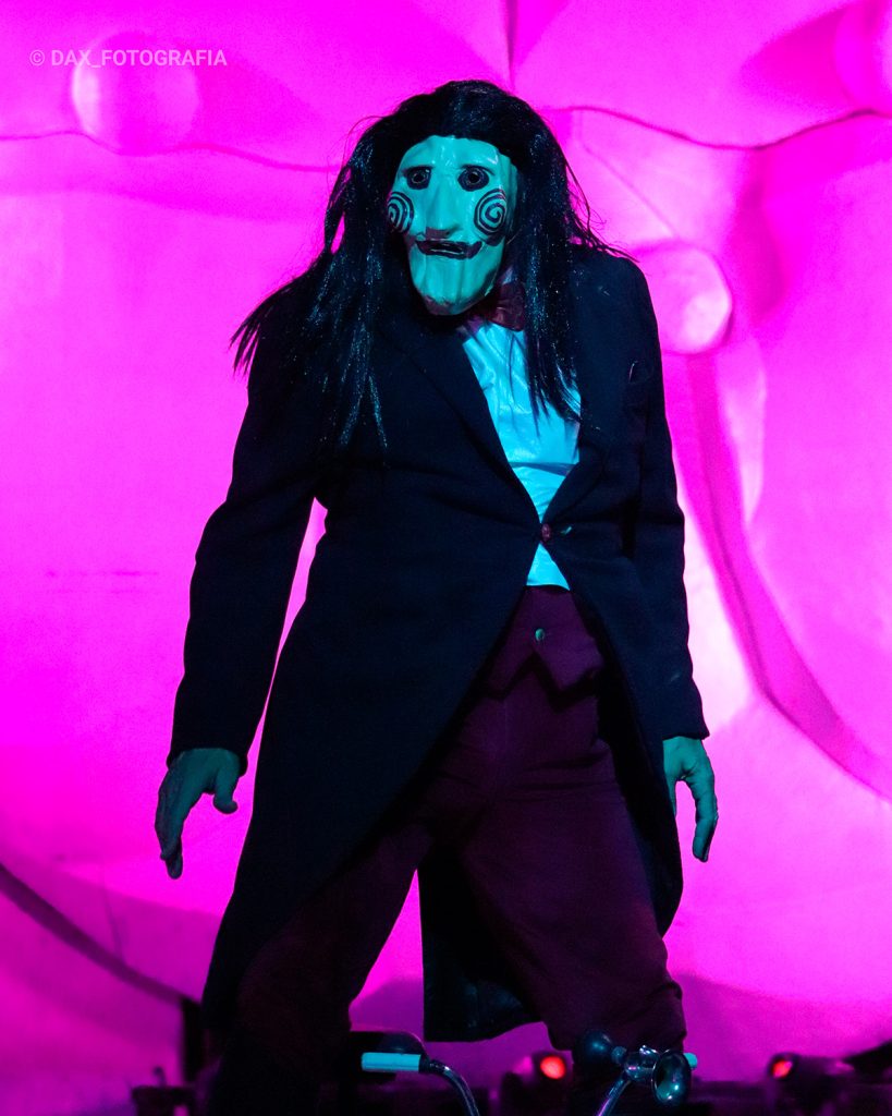 Intérprete con rostro pálido y traje negro aparece iluminado sobre un fondo magenta.