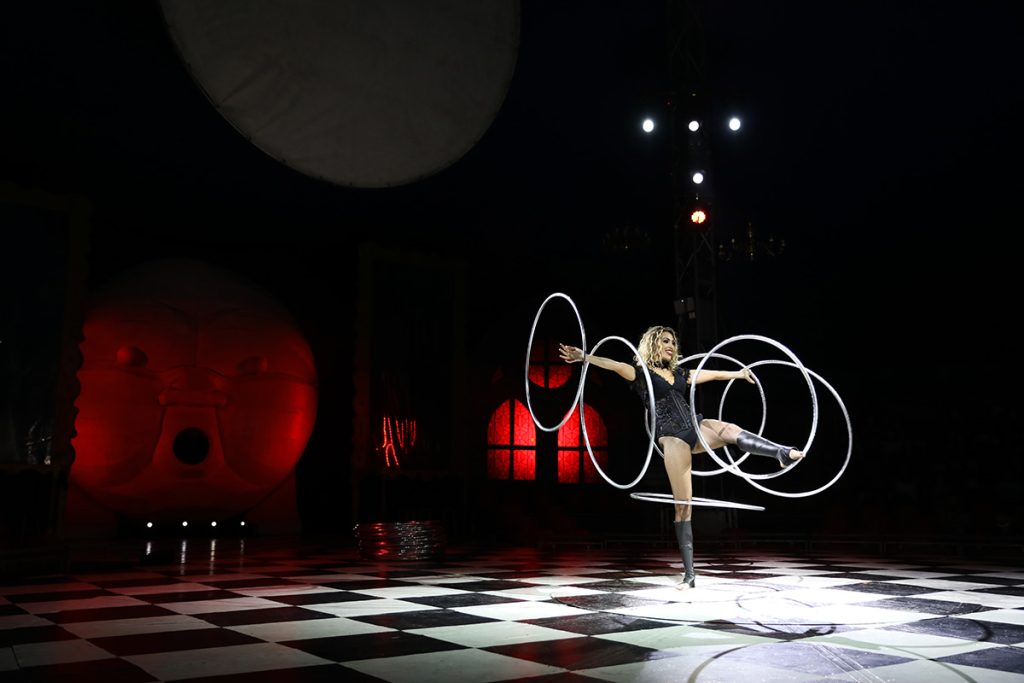 Pista de circo con suelo ajedrezado y varios aros frente a un fondo oscuro.