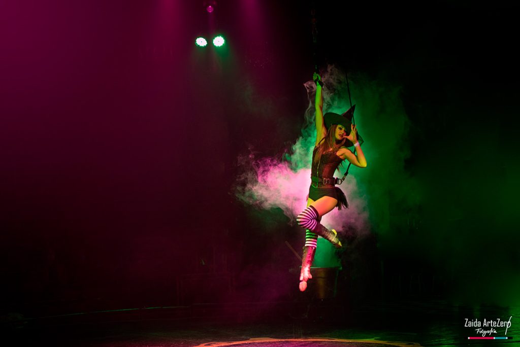 Artista de circo suspendida en el aire bajo un foco verde y entre niebla escénica.
