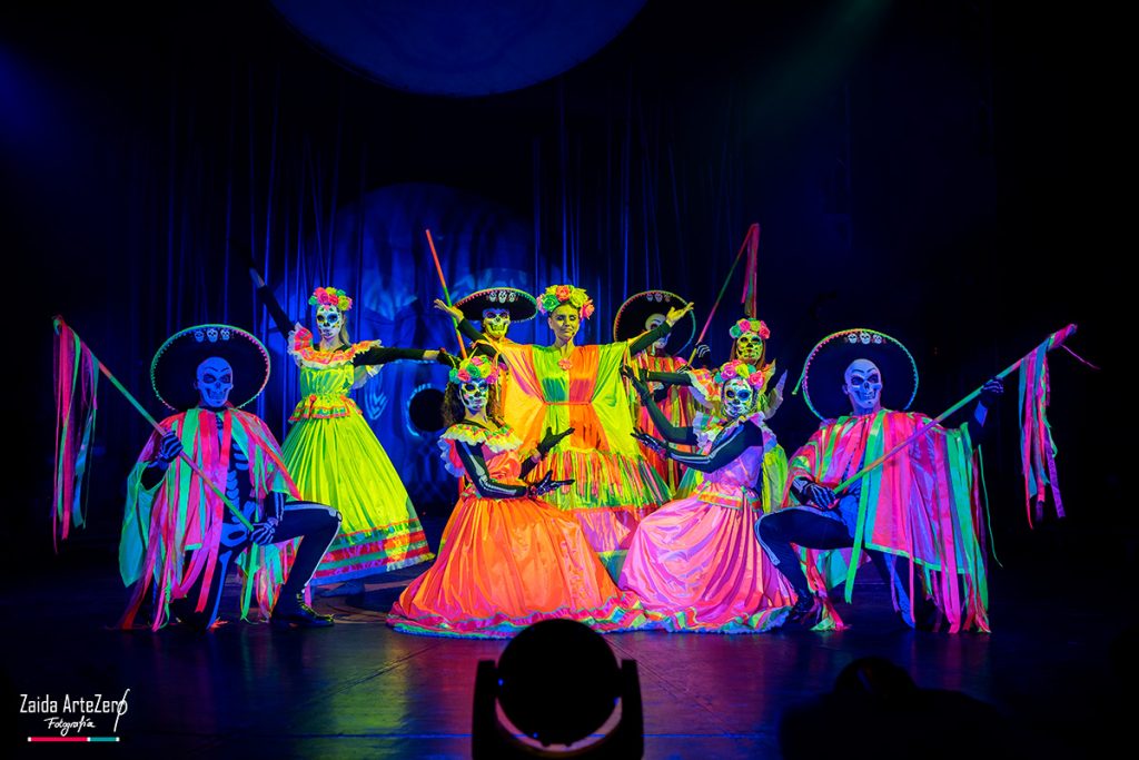 Varios artistas del espectáculo, con vestuario de colores, aparecen juntos en un escenario iluminado.