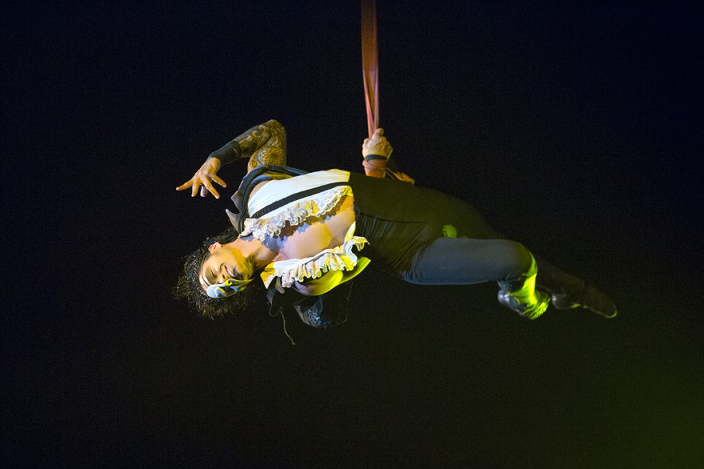Artista realiza una figura acrobática suspendida en el aire bajo un foco sobre fondo oscuro.