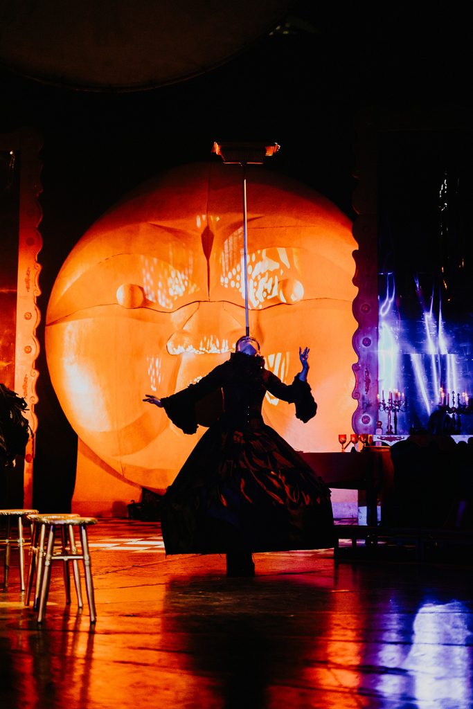 Silueta de una artista con vestido amplio ante un gran círculo naranja iluminado.
